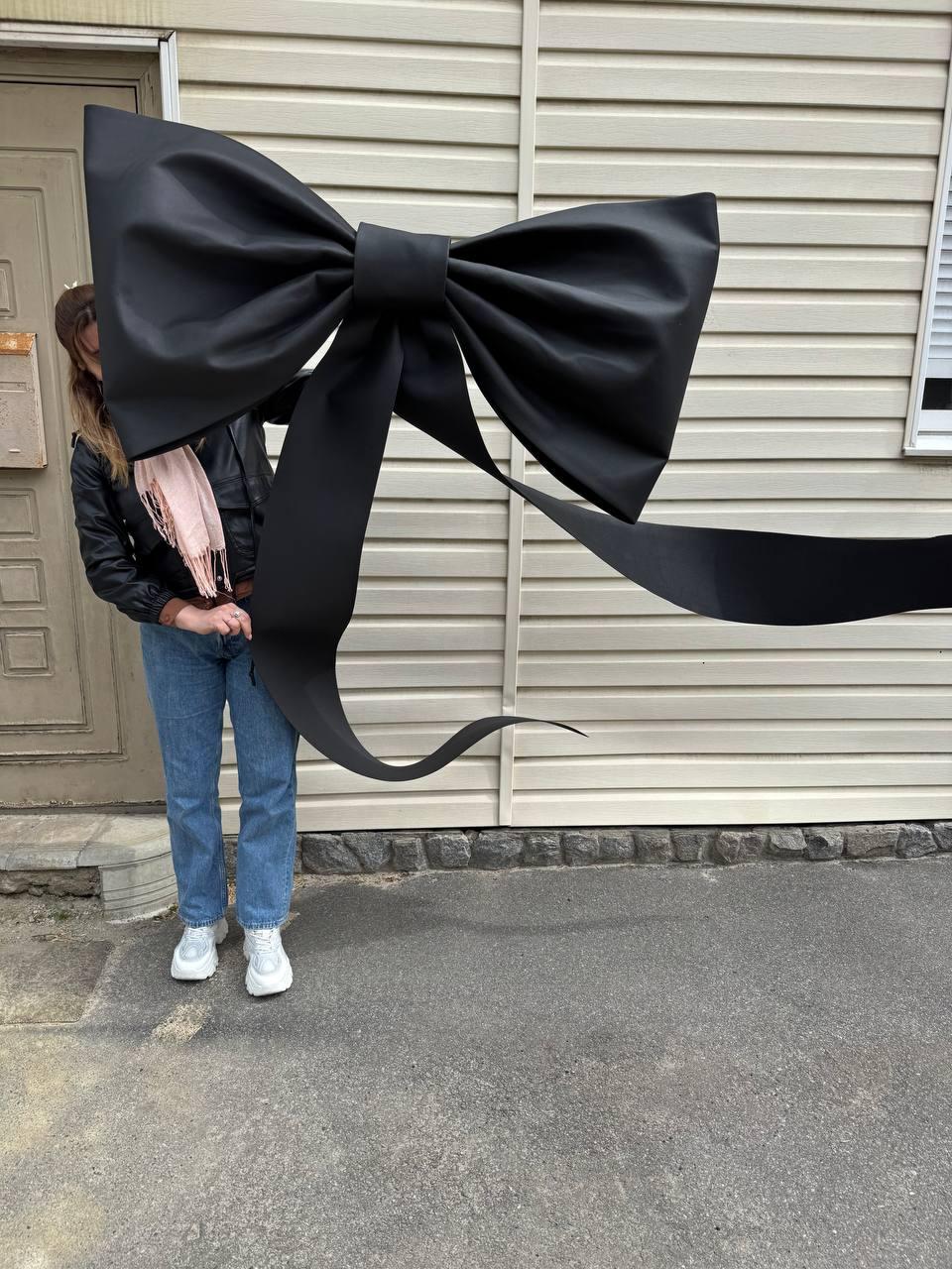May include: A large, black bow made of fabric. The bow is held up in front of a beige wall. The bow has long, flowing tails. The bow is a decorative item, possibly for a party or event.