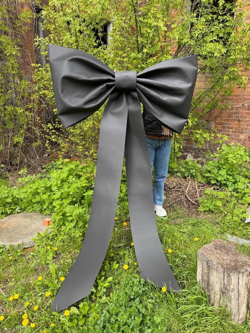 May include: A large, black bow with long, flowing tails stands in a grassy area with a brick building in the background. The bow is made of a smooth, matte material and is a striking decorative element.