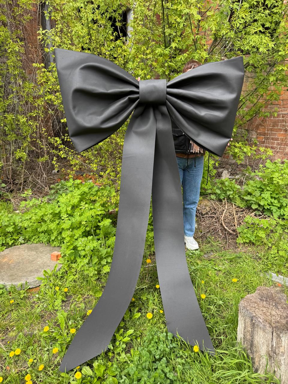 May include: A large, black bow with long, flowing tails. The bow is made of a matte material and is displayed outdoors against a backdrop of green foliage and a brick building. The bow's size suggests it could be used for decoration.