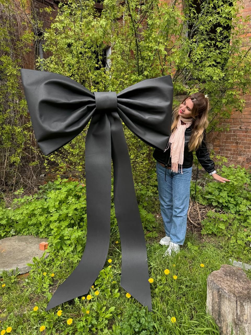 May include: A large, black bow with long, flowing tails stands in a grassy area. The bow is made of a smooth, possibly satin-like material. The bow is the focal point, set against a backdrop of green foliage and a brick building.