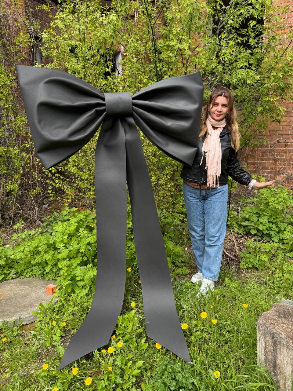 May include: A large, black bow with long, flowing tails stands in a grassy area. The bow is made of a matte material and has a classic bow shape. The background includes green foliage and a brick building.