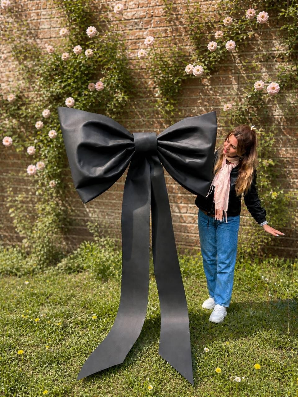May include: A large, black bow stands on a grassy lawn in front of a brick wall covered in pink flowers. The bow has long, flowing tails. The bow is a decorative element.