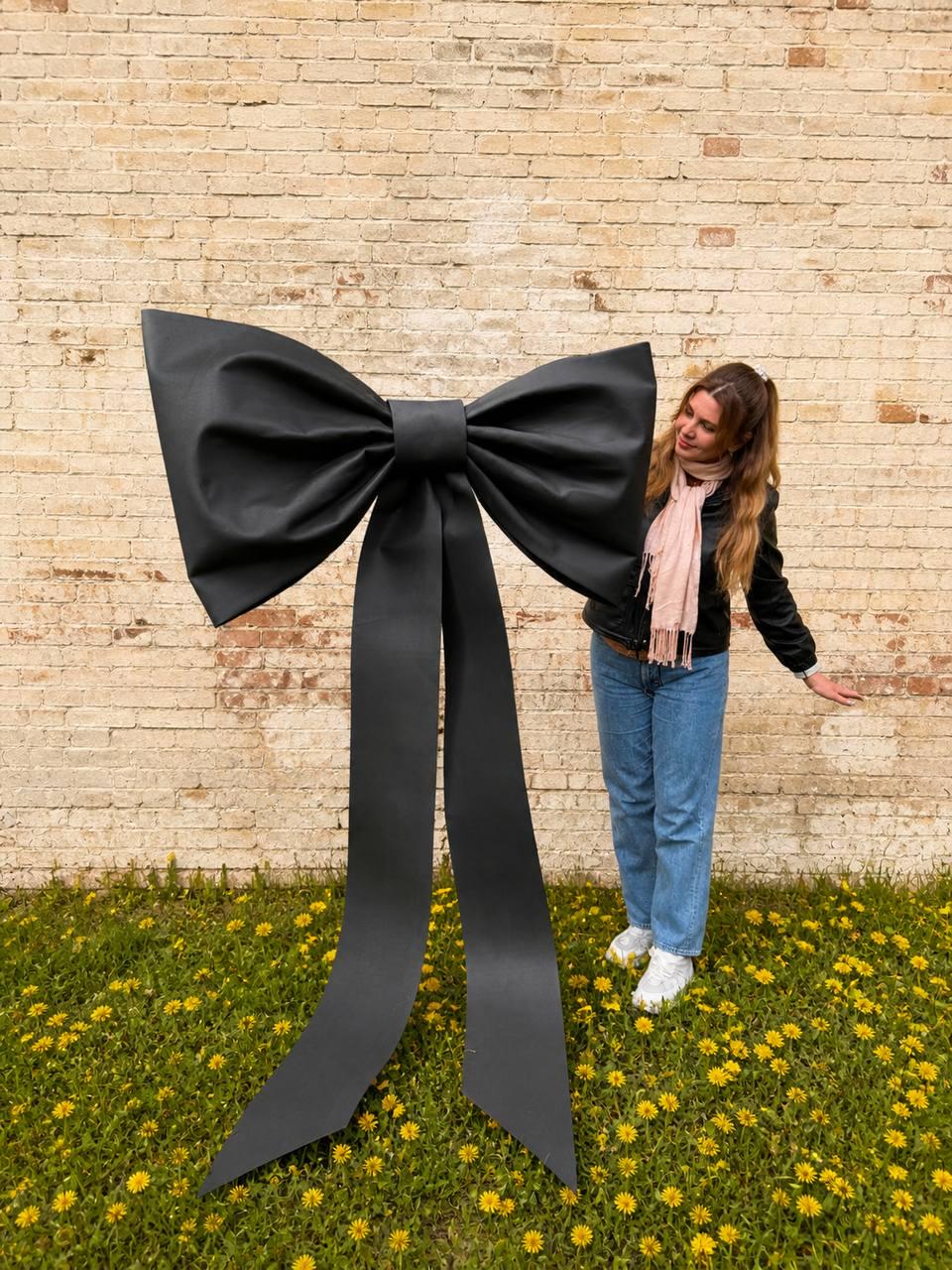 May include: A large, black bow with long, flowing tails stands in front of a brick wall. The bow is made of a smooth, matte material. The bow is positioned in a field of green grass and yellow flowers.