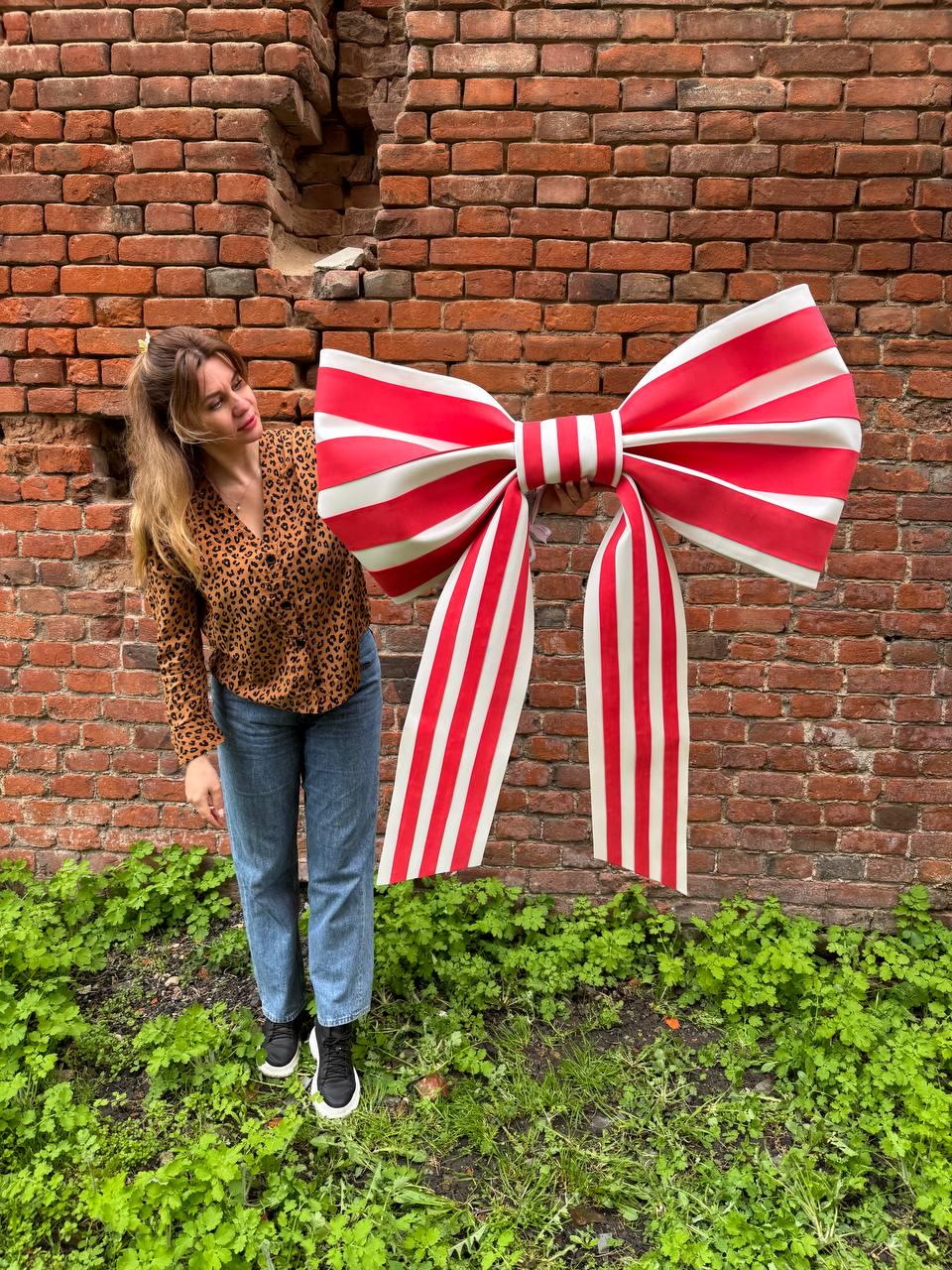May include: A large, decorative bow with red and white stripes. The bow is held in front of a brick wall and green foliage. The bow's dimensions are approximately 48 inches wide and 60 inches long.