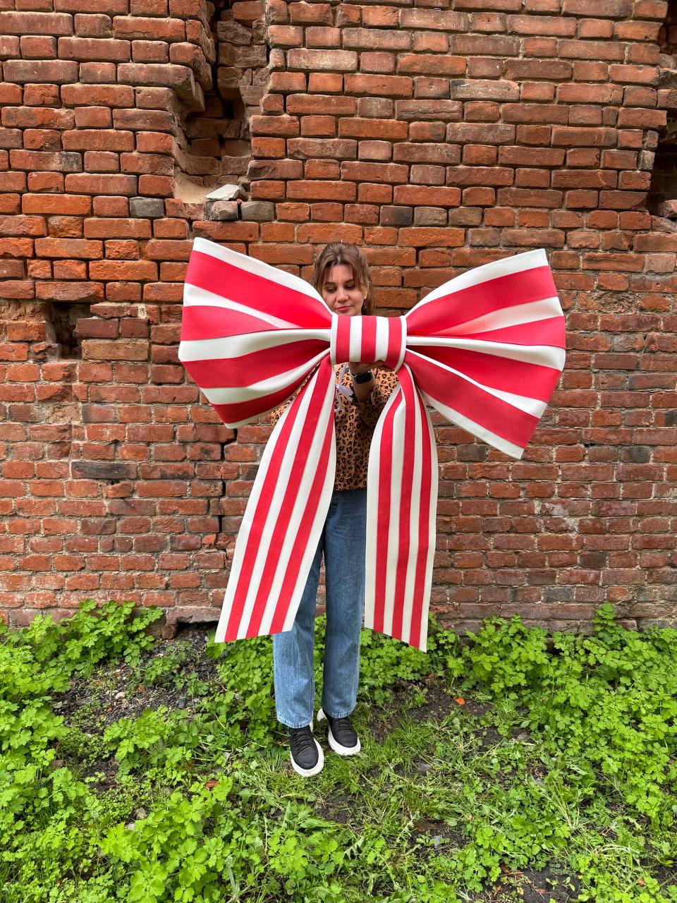 May include: A large, decorative bow with red and white stripes. The bow is held in front of a person, against a backdrop of a brick wall and green foliage. The bow's size is substantial, making it a striking visual element.