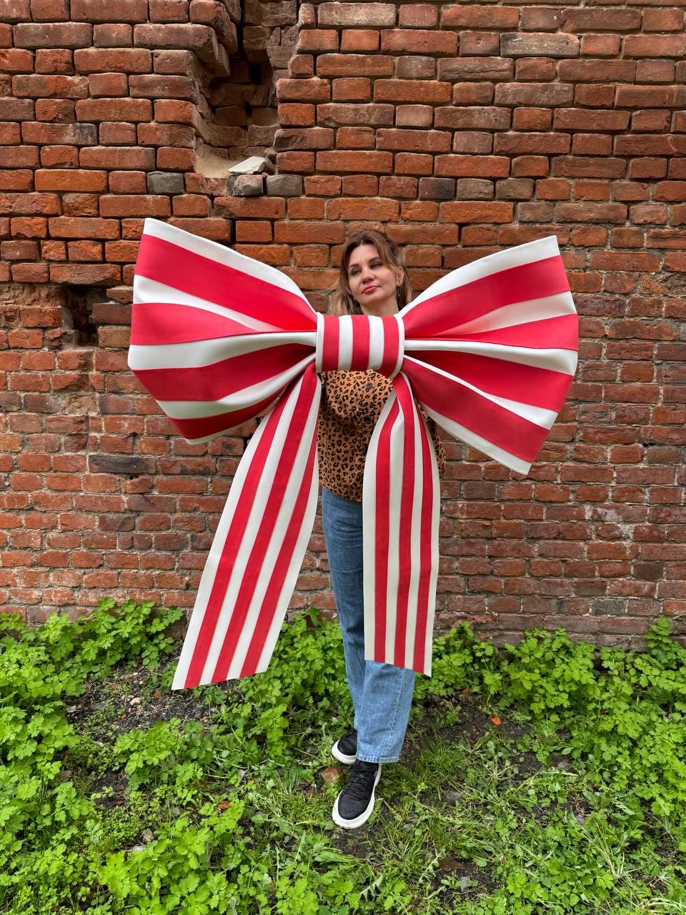May include: A large, decorative bow with red and white stripes. The bow is oversized and appears to be made of a sturdy material. The bow is in front of a brick wall and green foliage.