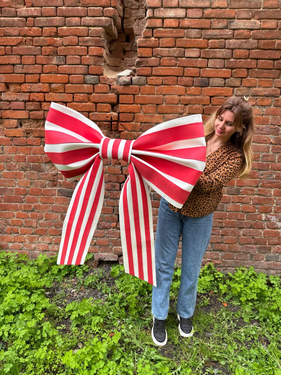 May include: A large, decorative bow with red and white stripes. The bow is held in front of a brick wall and green foliage. The bow's design features a classic striped pattern, making it suitable for festive occasions or decorative purposes.