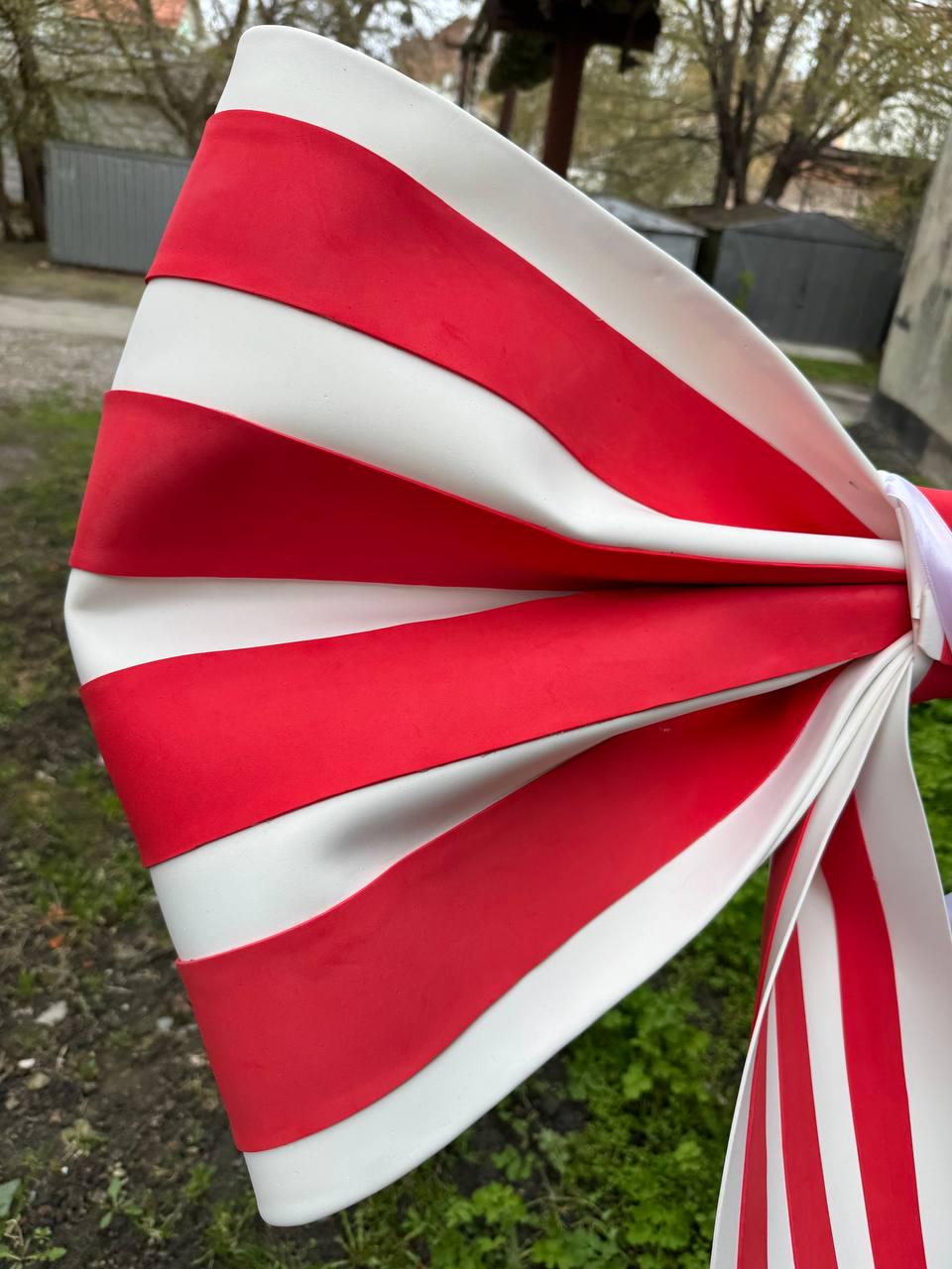 May include: A large, decorative bow with alternating red and white stripes. The bow is made of a flexible material and is tied with a white ribbon. The bow is outdoors, with green grass and a building in the background.
