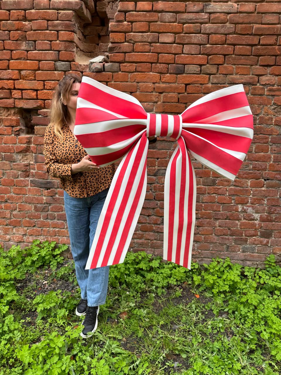 May include: A large, decorative bow with red and white stripes. The bow is held in front of a brick wall and green foliage. The bow's design features a central knot with long, striped ribbons extending downwards. The bow is a festive decoration.