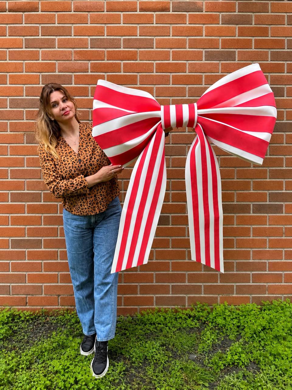 May include: A large, decorative bow with red and white stripes. The bow is held in front of a brick wall and has long, striped streamers. The bow is a festive decoration, possibly for a party or event.