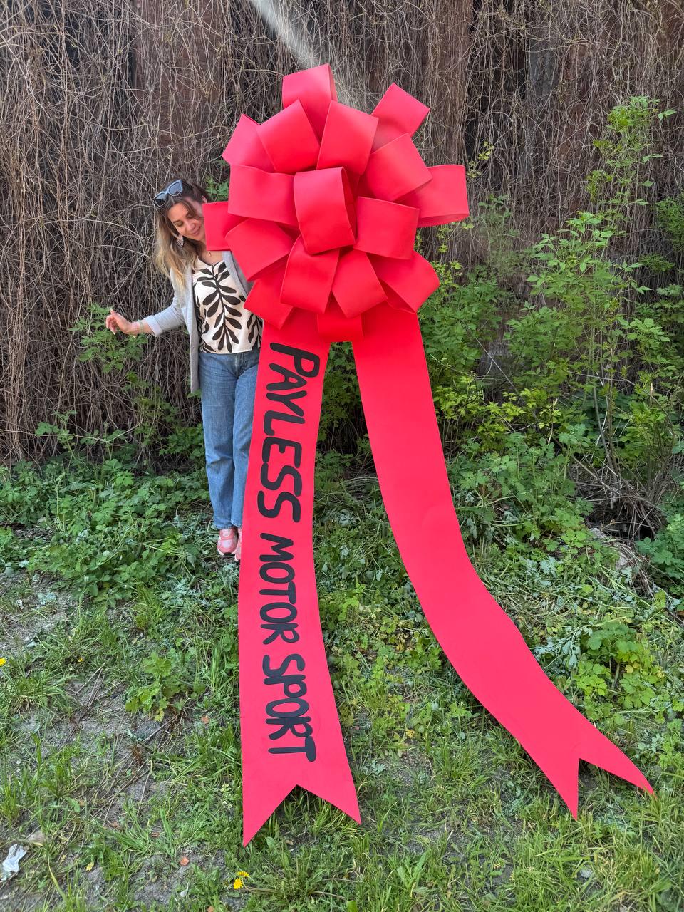 May include: A large, bright red bow with long, flowing ribbons. The bow has the text "PAYLESS MOTOR SPORT" printed in black on one of the ribbons. The bow is set against a backdrop of green foliage and a person.