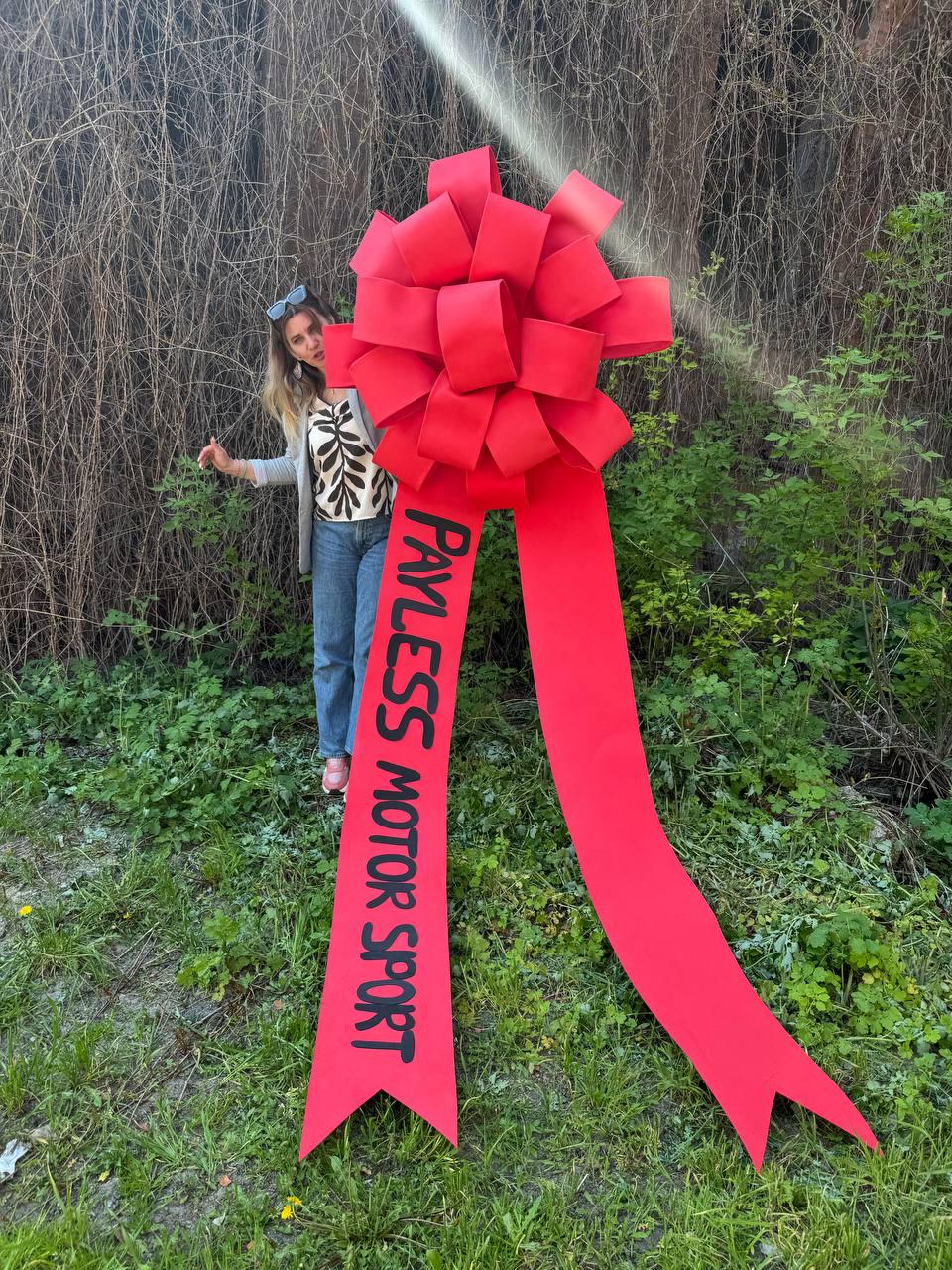 May include: A large red bow with long ribbons, the text "PAYLESS MOTOR SPORT" is printed in black on one of the ribbons. The bow is in front of a person standing in a grassy area with a backdrop of bushes and trees.