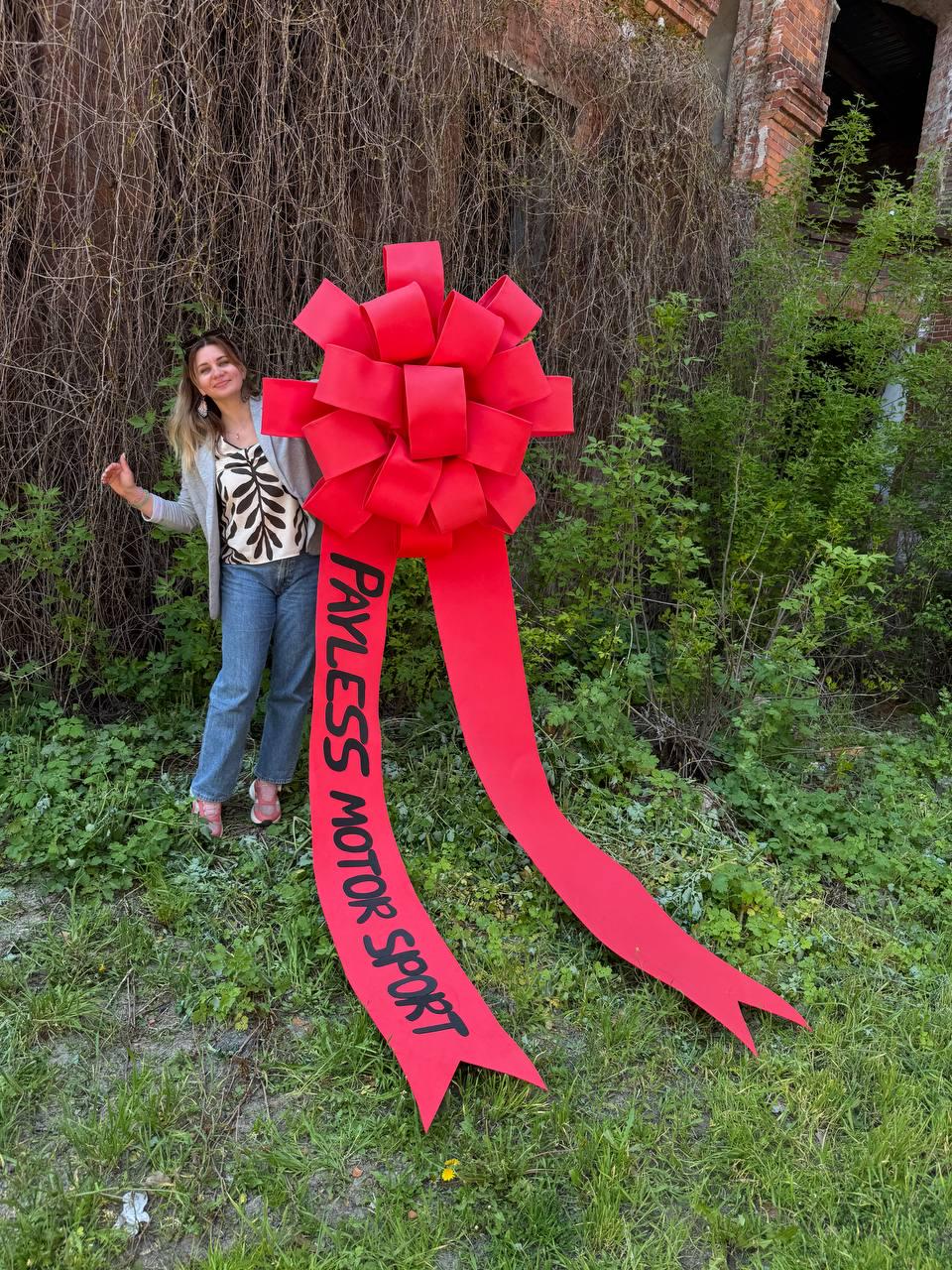 May include: A large, bright red bow with long streamers. The bow has the text "PAYLESS MOTOR SPORT" printed in black. The bow is displayed outdoors with a person standing next to it.