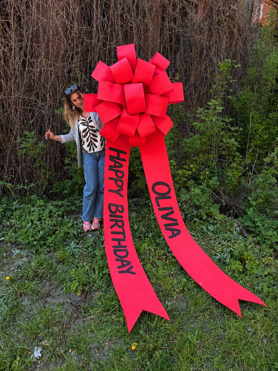May include: A large, bright red bow with long, flowing ribbons. The ribbons read "HAPPY BIRTHDAY OLIVIA" in black text. The bow is set up outdoors in front of greenery.