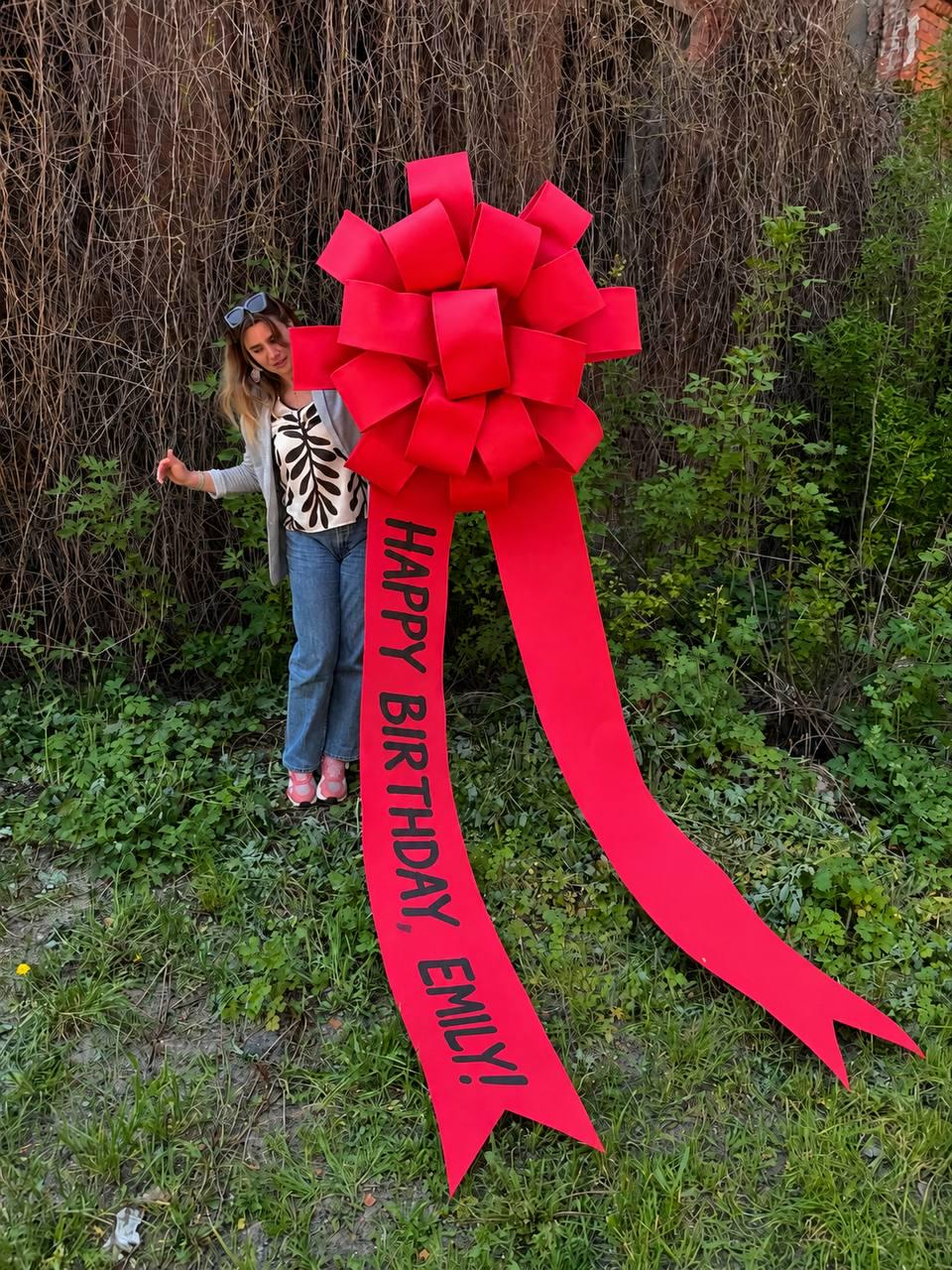 May include: A large, bright red bow with long streamers. The bow has the text "HAPPY BIRTHDAY, EMILY!" printed in black. The bow is set outdoors in front of greenery.
