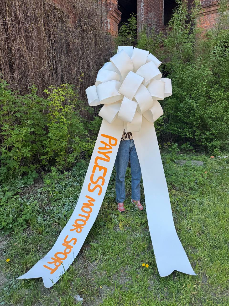 May include: A large white bow with long streamers, featuring the text "PAYLESS MOTOR SPORT" in orange. The bow is set against a backdrop of green foliage and an old brick building.