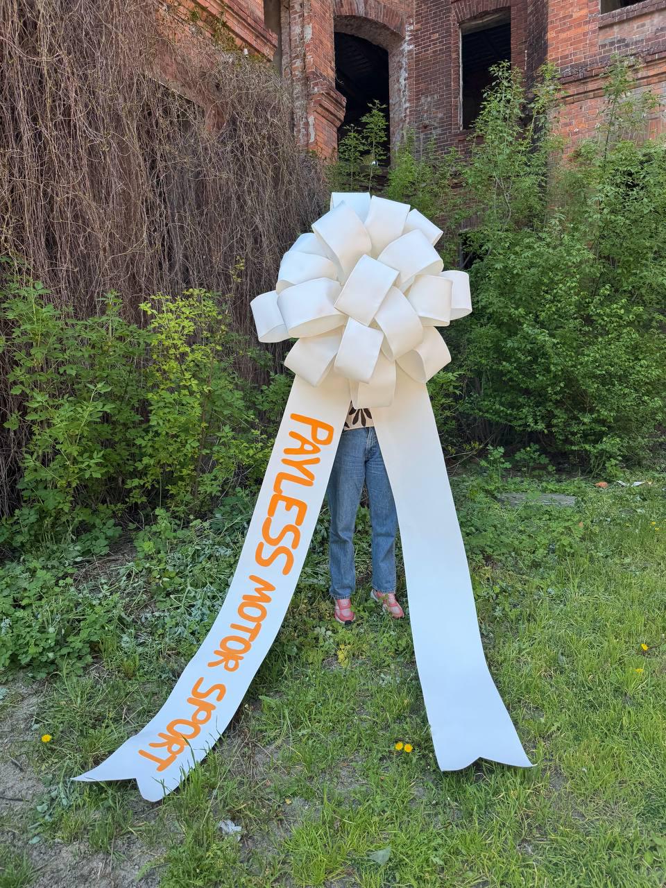 May include: A large white bow with long streamers. The streamers have the words "PAYLESS MOTOR SPORT" printed in orange. The bow is in front of a person standing on grass, with a brick building in the background.