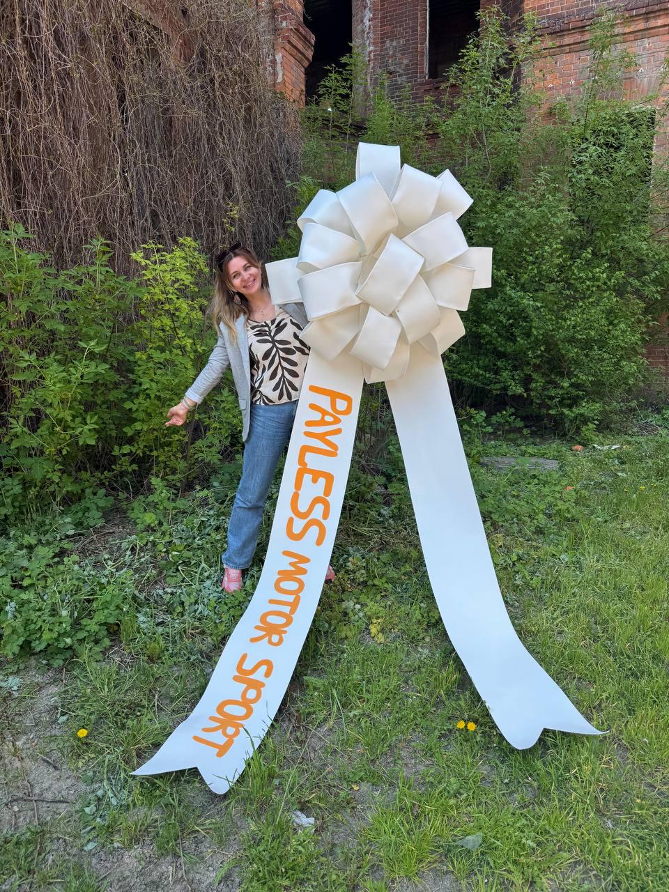 May include: A large, white bow with long streamers, featuring the text "PAYLESS MOTOR SPORT" in orange. The bow is positioned outdoors with a person standing nearby. The background includes greenery and a brick building.