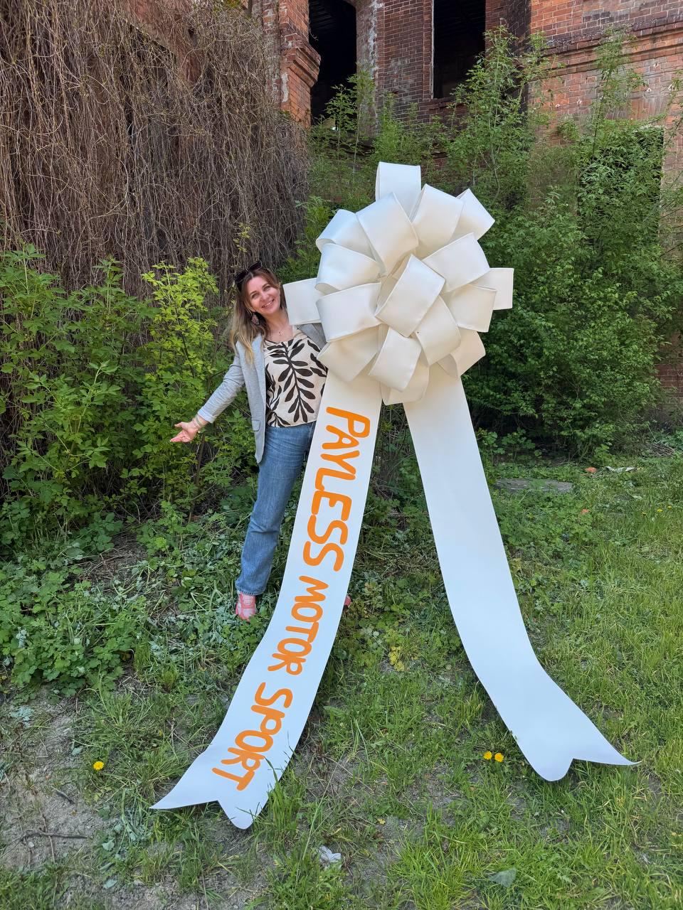 May include: A large, white bow with long streamers, featuring the text "PAYLESS MOTOR SPORT" in orange. The bow is positioned outdoors with a person standing nearby. The background includes greenery and a brick building.