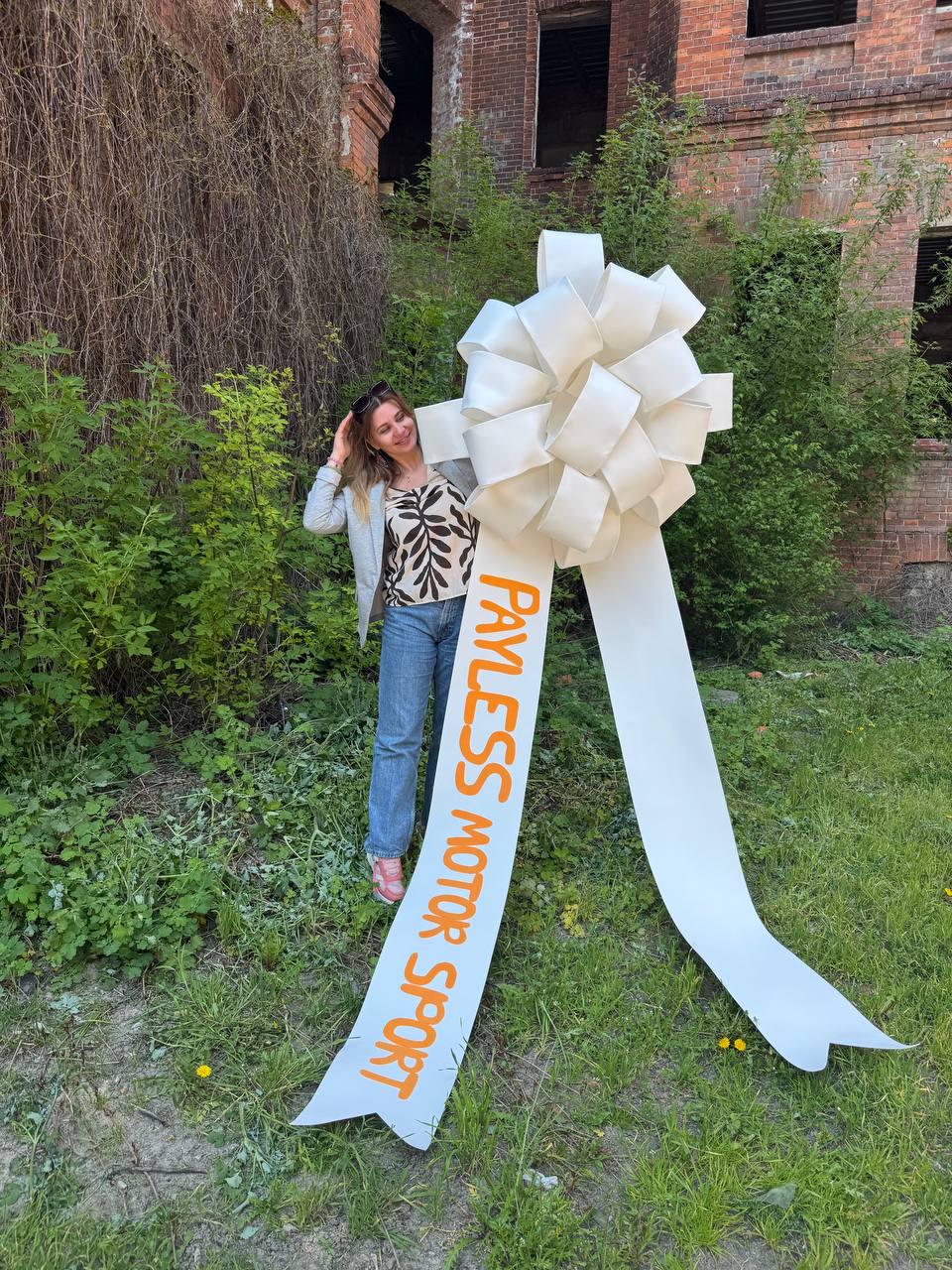 May include: A large white bow with long streamers. The text "PAYLESS MOTOR SPORT" is printed in orange on one of the streamers. The bow is in front of a person wearing a patterned top, jeans, and a blazer.