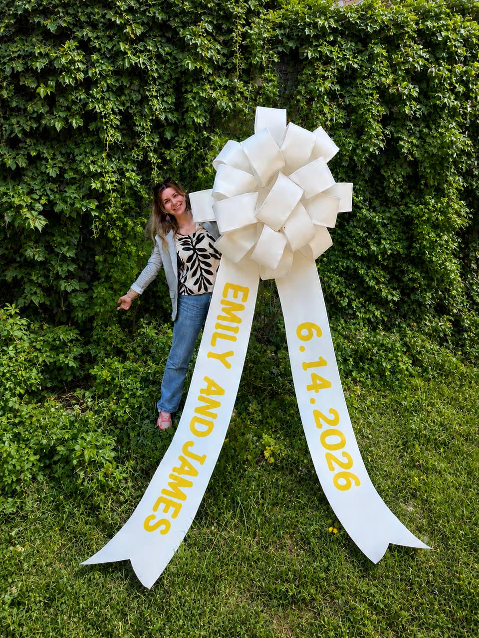 May include: A large white bow with long ribbons, featuring the text "EMILY AND JAMES" and the date "6.14.2026" in yellow. The bow is set against a backdrop of lush green foliage and grass.