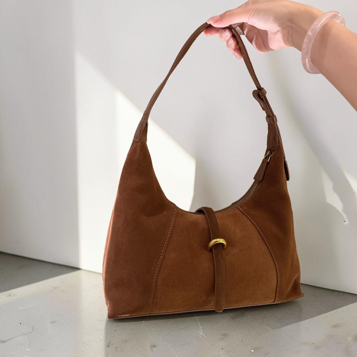 May include: A brown suede shoulder bag with a curved design. The bag features a long strap, a gold-colored buckle, and a zipper closure. The bag is held by a hand.
