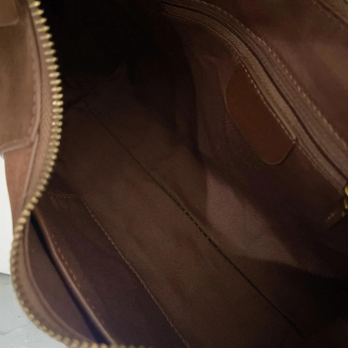 May include: Open view of a brown leather handbag interior. The bag features a brown fabric lining, a small pocket, and a gold-colored zipper. The bag is designed with a classic, minimalist style.