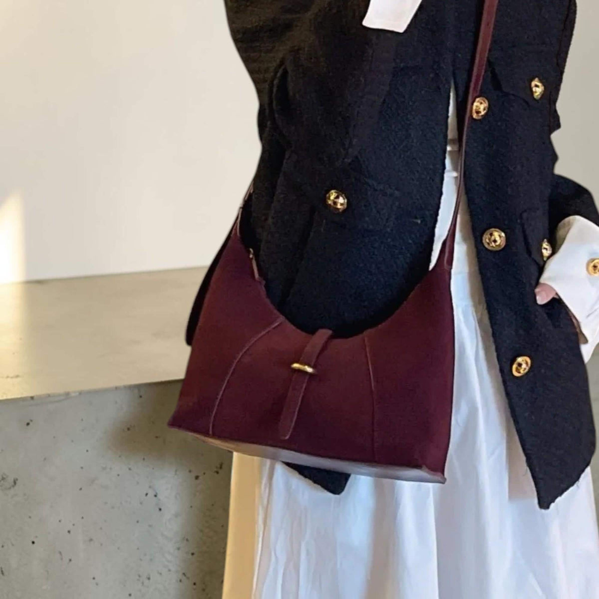 May include: A burgundy shoulder bag with a curved design and a gold-colored buckle. The bag has a long strap and is made of a soft material. The bag is worn over a black jacket with gold buttons and a white dress.