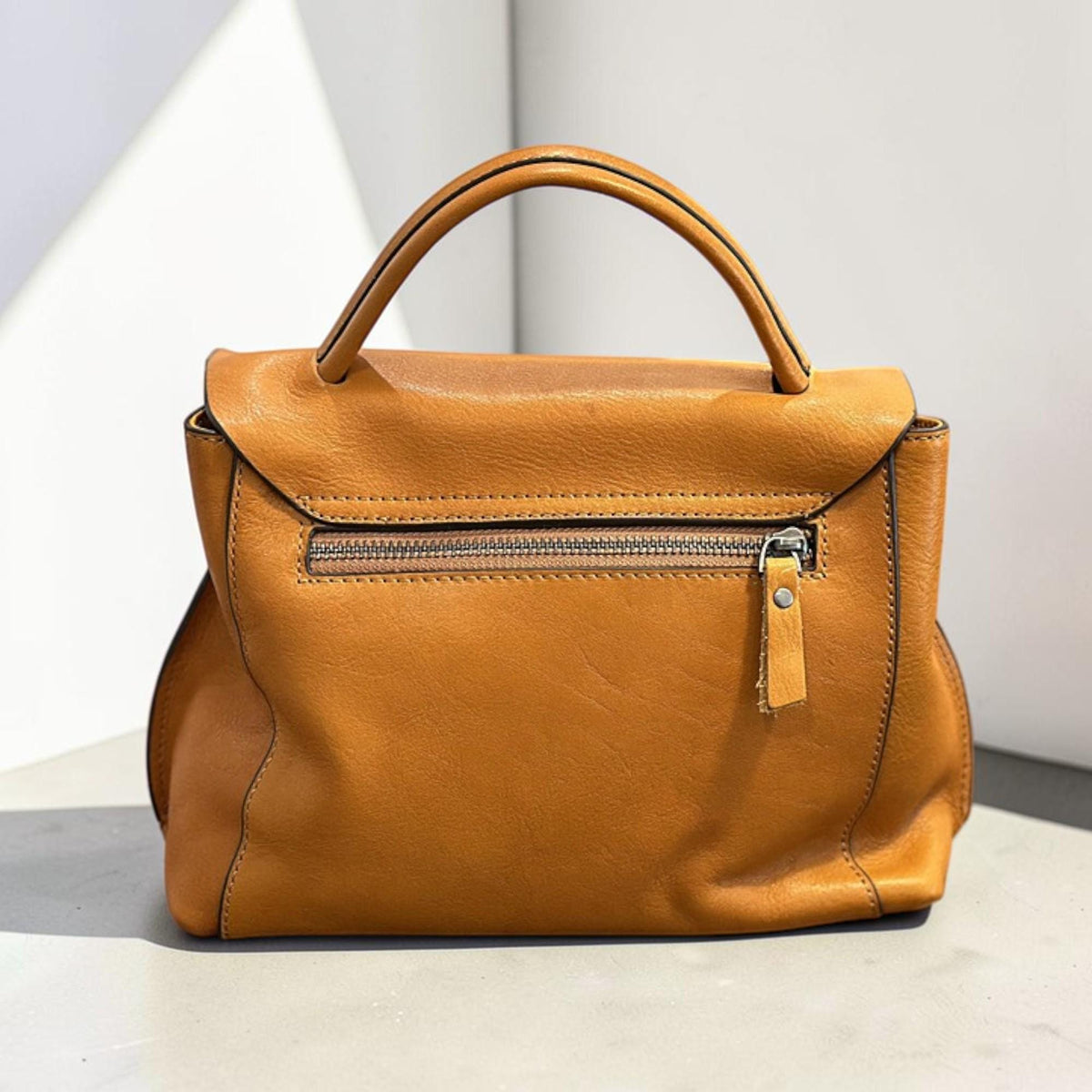 May include: A tan leather handbag with a top handle and a flap closure. The bag features a zipper pocket on the back and a small leather pull tab. The stitching is visible along the edges, and the bag appears to be made of high-quality leather.