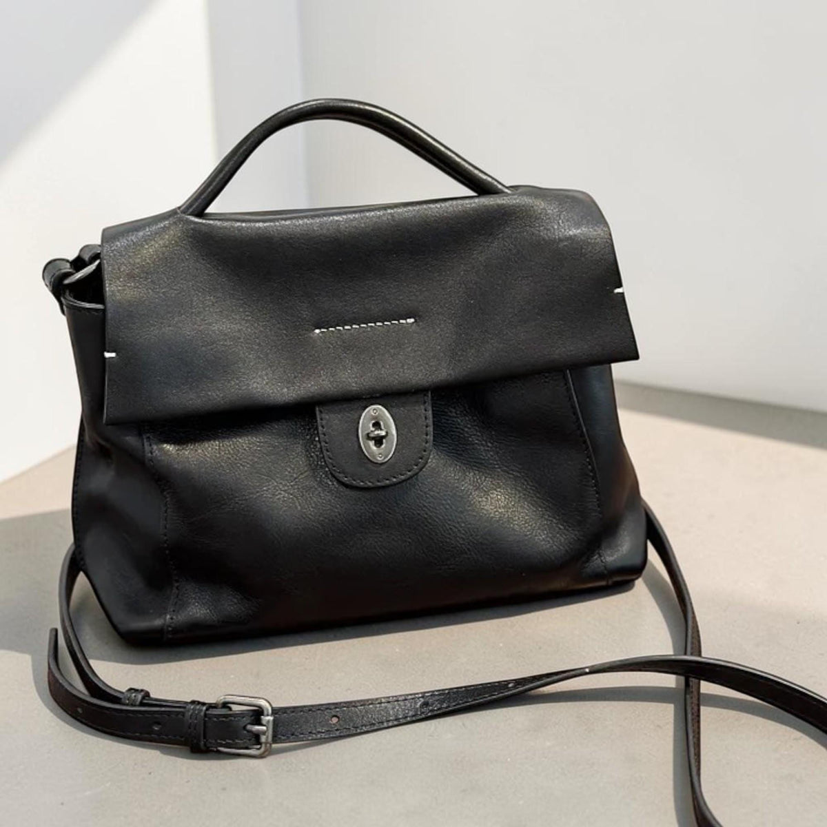May include: A black leather handbag with a top handle and a detachable shoulder strap. The bag features a flap closure with a silver-toned clasp. The design includes a small top handle and a long, adjustable strap for crossbody wear.
