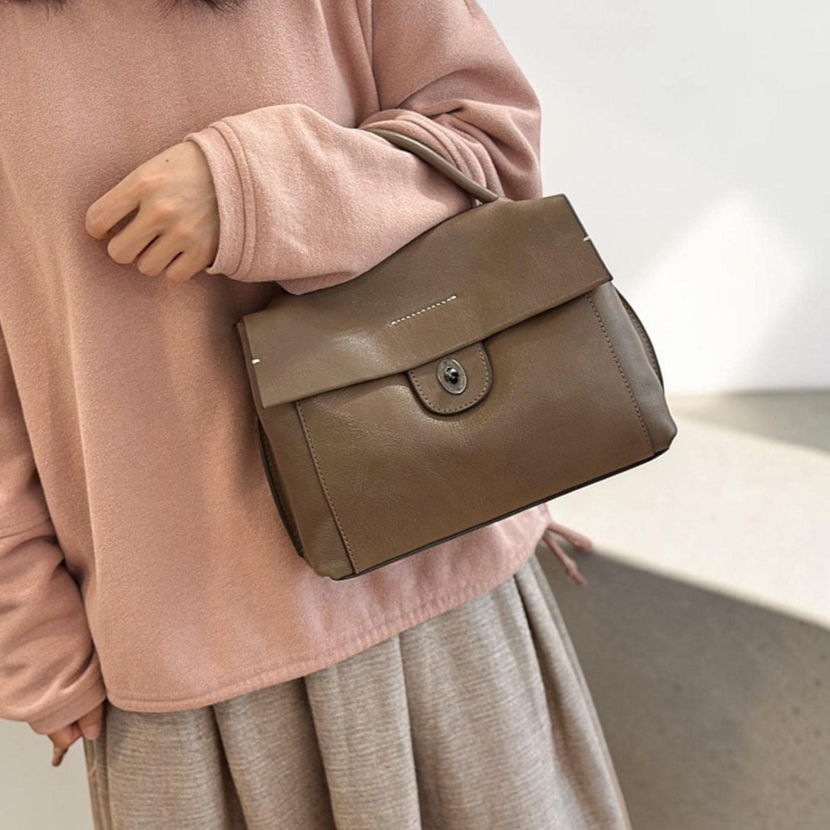 May include: A taupe-colored handbag with a flap closure and a small handle. The bag has a metal clasp and is made of a smooth material. The bag is being held by a person wearing a pink sweater and a gray skirt.