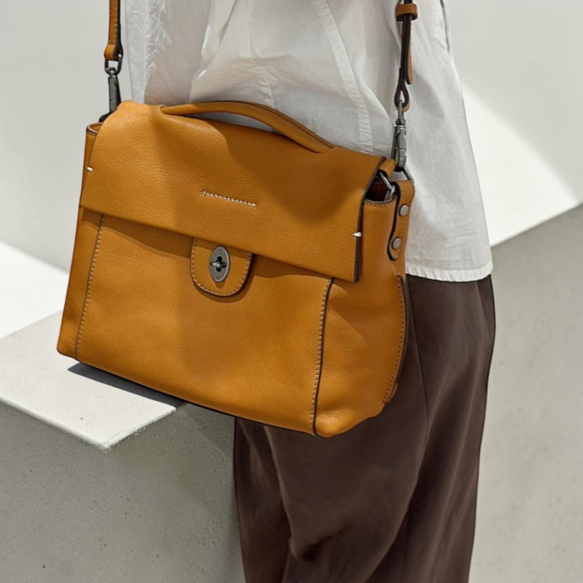 May include: A mustard-colored leather shoulder bag with a top handle and a flap closure. The bag has a rectangular shape with a silver-tone clasp and a long, adjustable strap. The bag is worn over the shoulder.