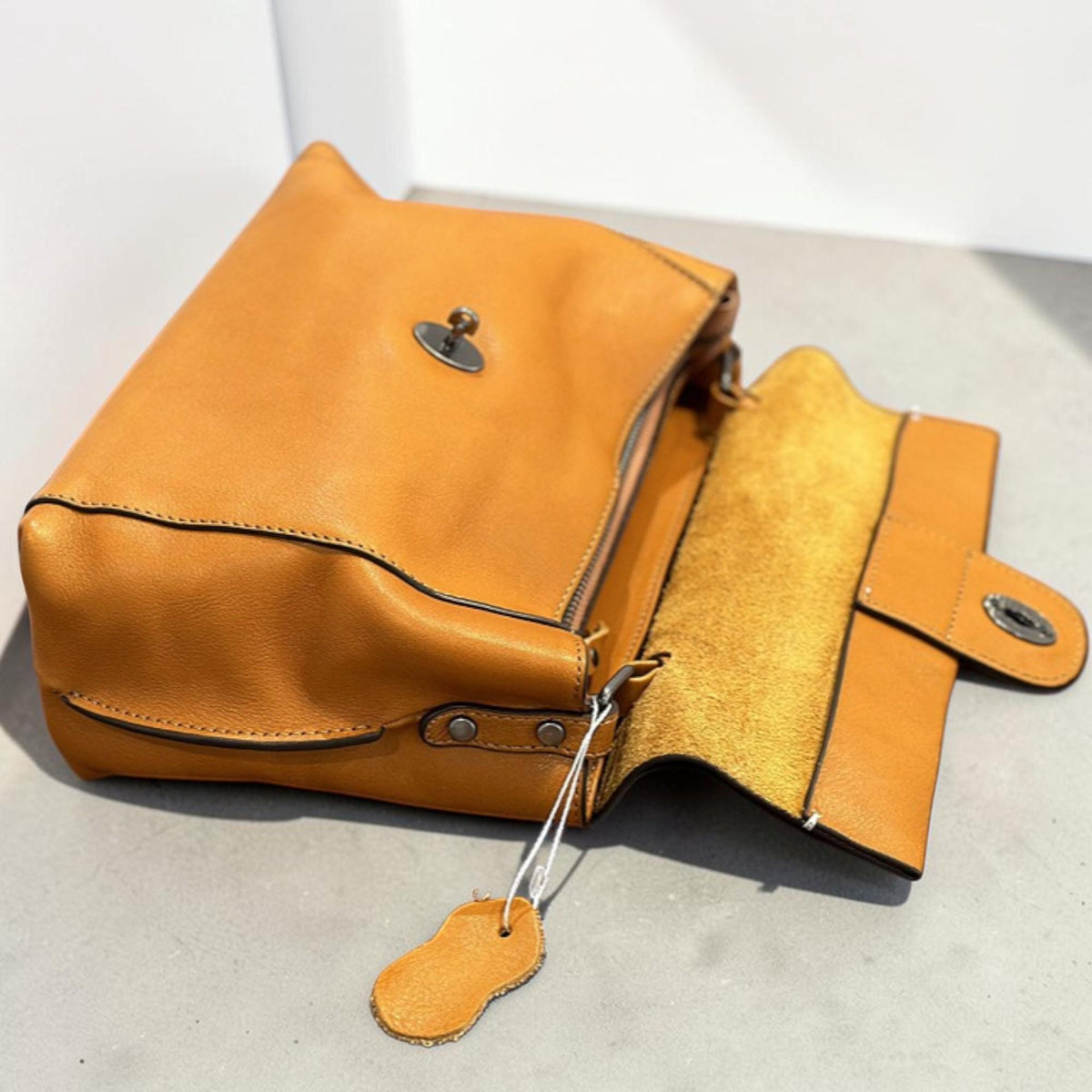 May include: A mustard-colored leather crossbody bag with a flap closure and a small tag. The bag is open, revealing a lighter interior. The bag has a metal clasp and stitching details.
