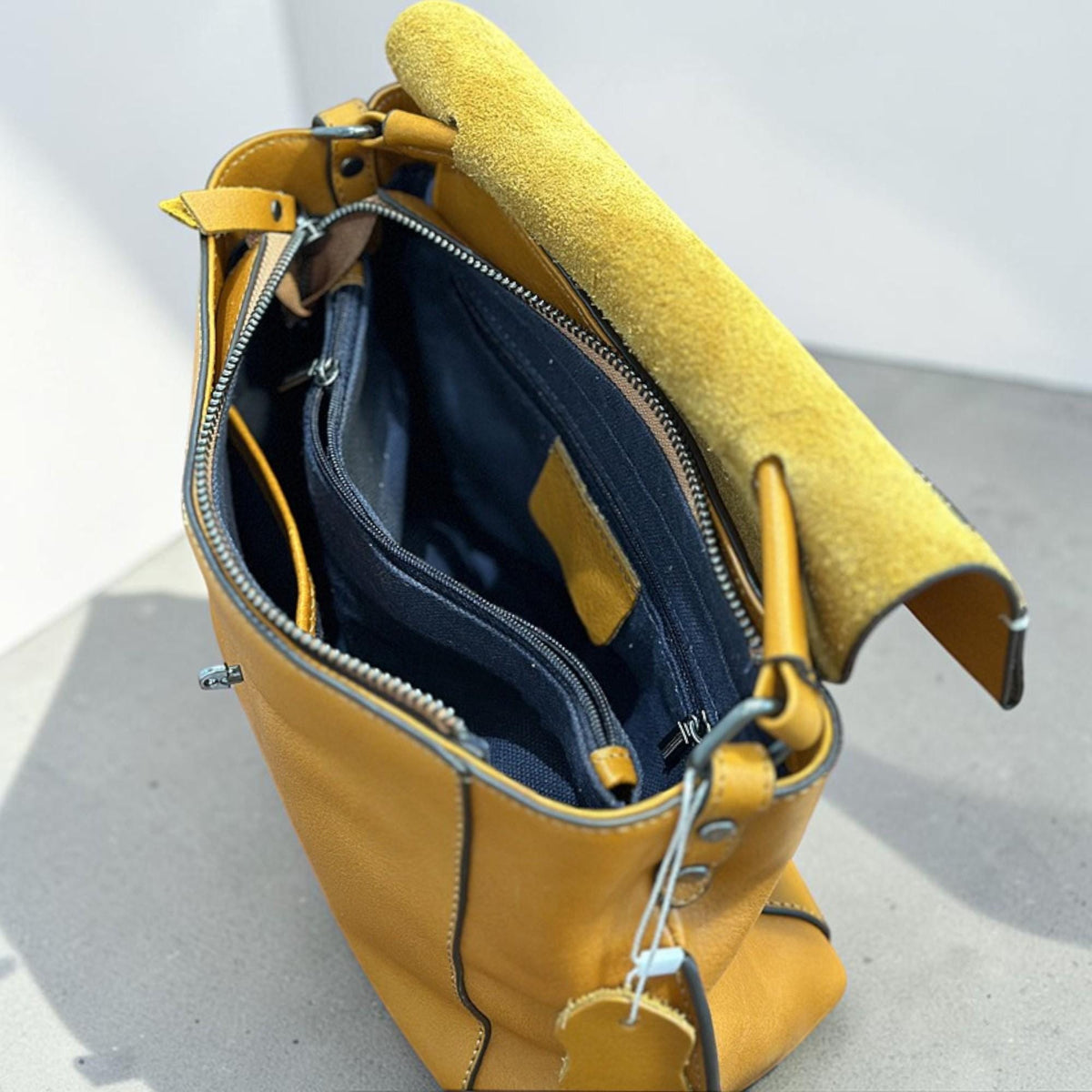 May include: A mustard yellow leather handbag with an open flap, revealing a black interior with multiple compartments. The bag has a zipper closure and a suede-like flap. The bag has a tag attached.
