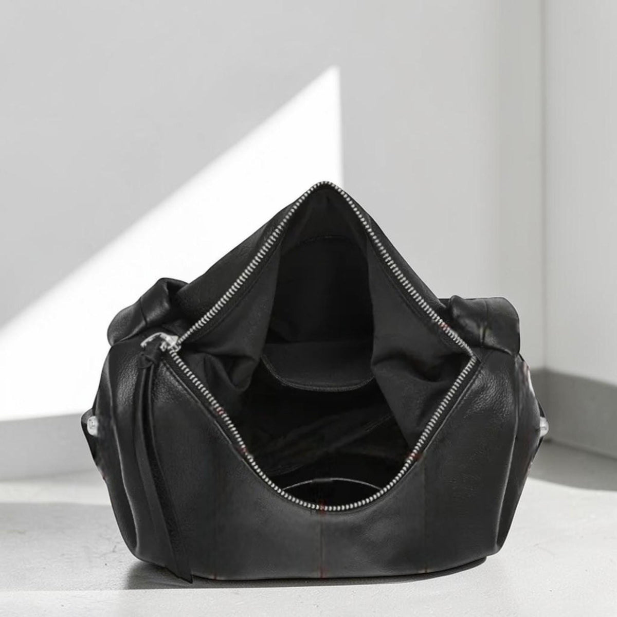 May include: Black leather handbag with a silver zipper, open to reveal the interior. The bag has a rounded shape and a minimalist design, suitable for everyday use.