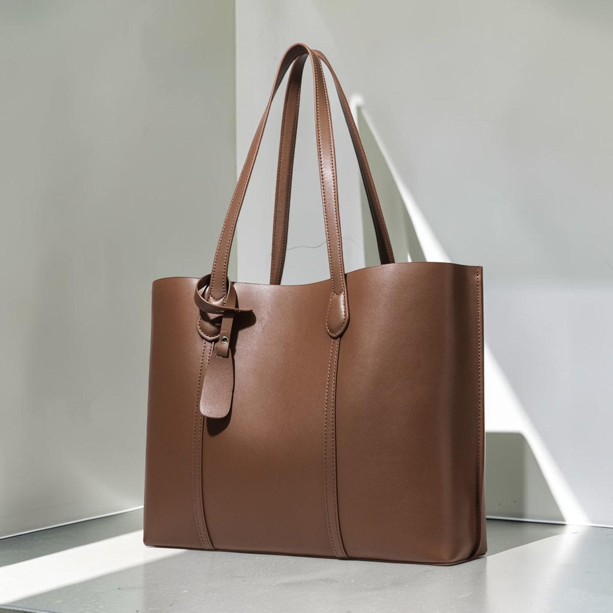 May include: A structured brown tote bag with two long shoulder straps, a smooth finish, and a decorative hanging leather tag attached to one of the handles. The bag features vertical stitched detailing on the front panel and a clean, open top design.