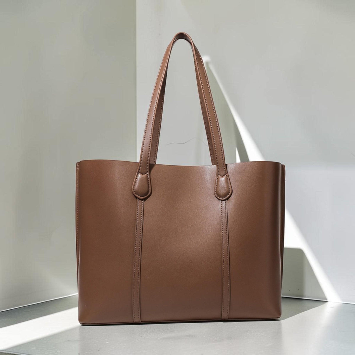 May include: A solid brown leather tote bag featuring a rectangular silhouette, two long shoulder straps attached with stitched detailing, and vertical seams running down the front panel, providing a clean and structured appearance for daily use.