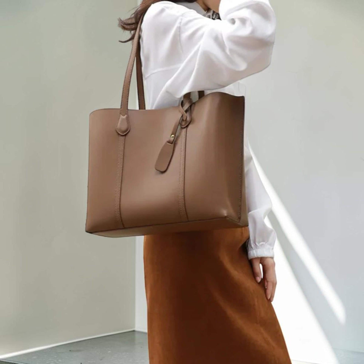 May include: A structured taupe tote bag with two vertical stitched panels on the front, thin shoulder straps, and a small dangling decorative tab attached to one side. The bag features a smooth, matte finish and a clean, rectangular silhouette.
