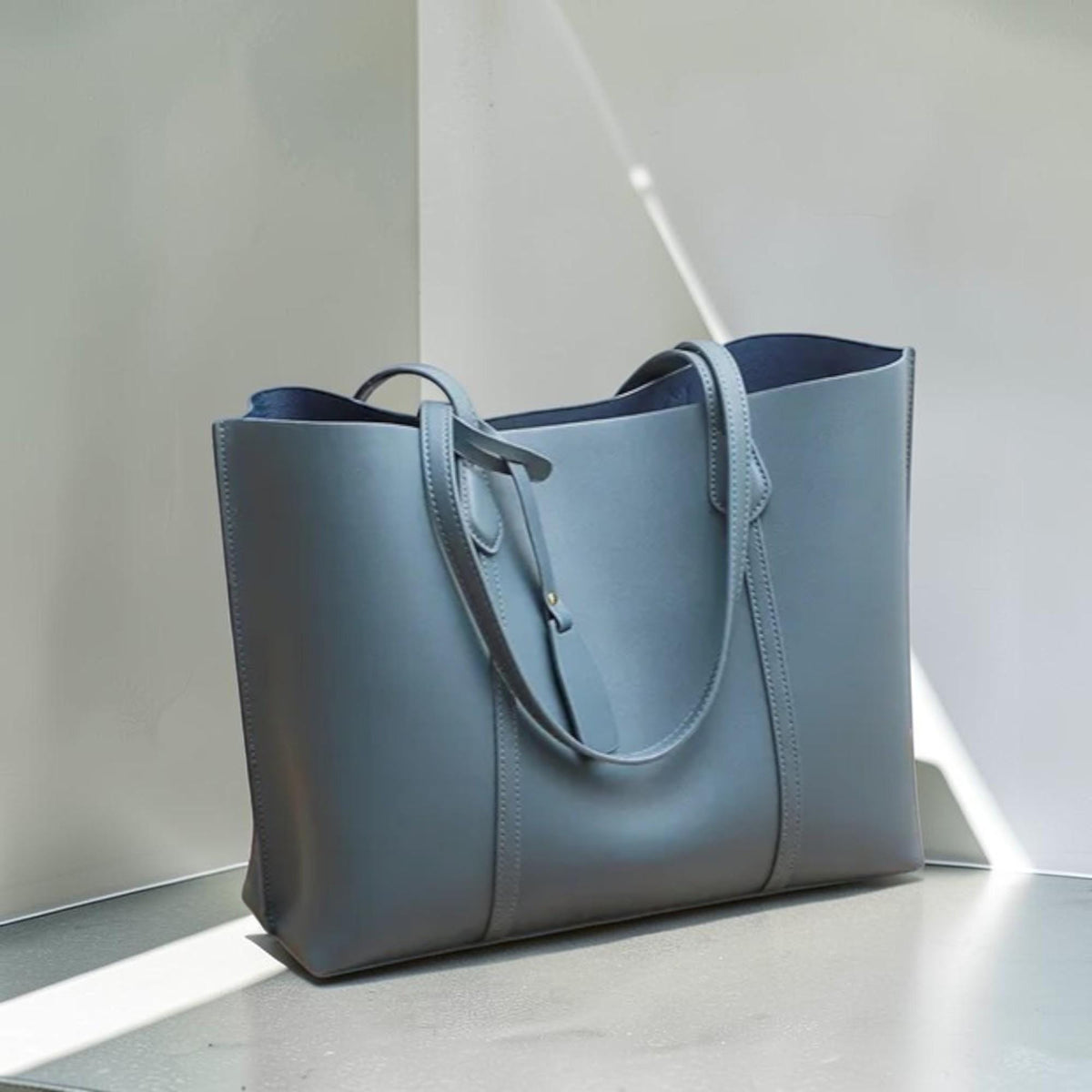May include: A slate blue tote bag with a structured rectangular silhouette, featuring two long shoulder straps, vertical stitched detailing on the front panel, and a small matching leather-like tag hanging from one of the handle attachments.