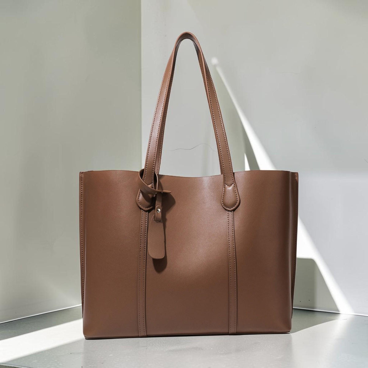 May include: A solid brown tote bag with a smooth finish, featuring two long shoulder straps and a decorative hanging leather-like tab attached to one strap with a small silver-toned metal rivet. The bag has a rectangular shape and vertical seams.