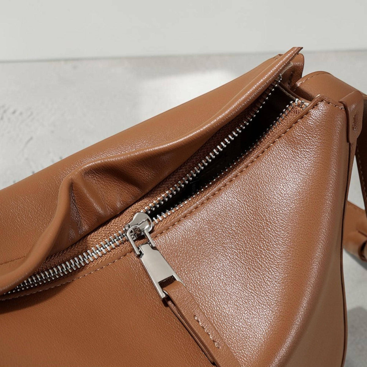 May include: Close-up of a brown leather bag with a silver zipper. The bag has a smooth texture and a rectangular shape. The zipper is partially open, revealing the interior of the bag.