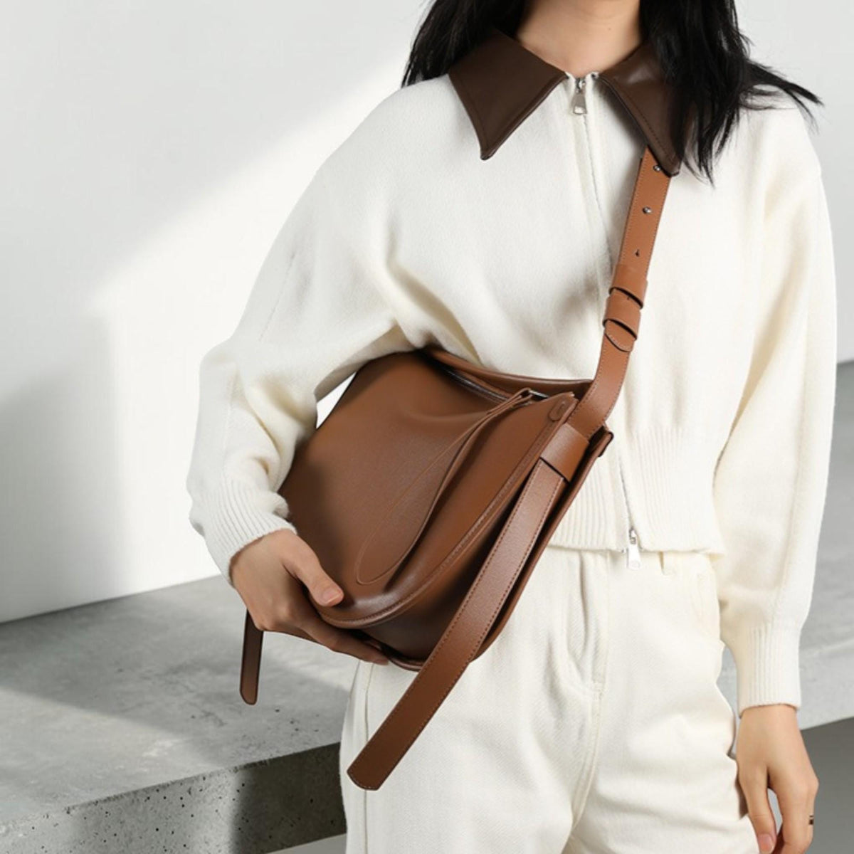 May include: A brown leather shoulder bag with a long strap. The bag has a rectangular shape with a curved flap. The model is wearing a white sweater with a brown collar and white pants.