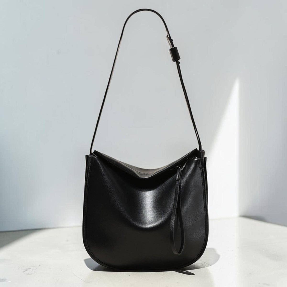 May include: A black leather shoulder bag with a curved, minimalist design. The bag has a long, thin strap and a small, attached handle. The bag is made of a smooth, shiny material and is set against a white background.
