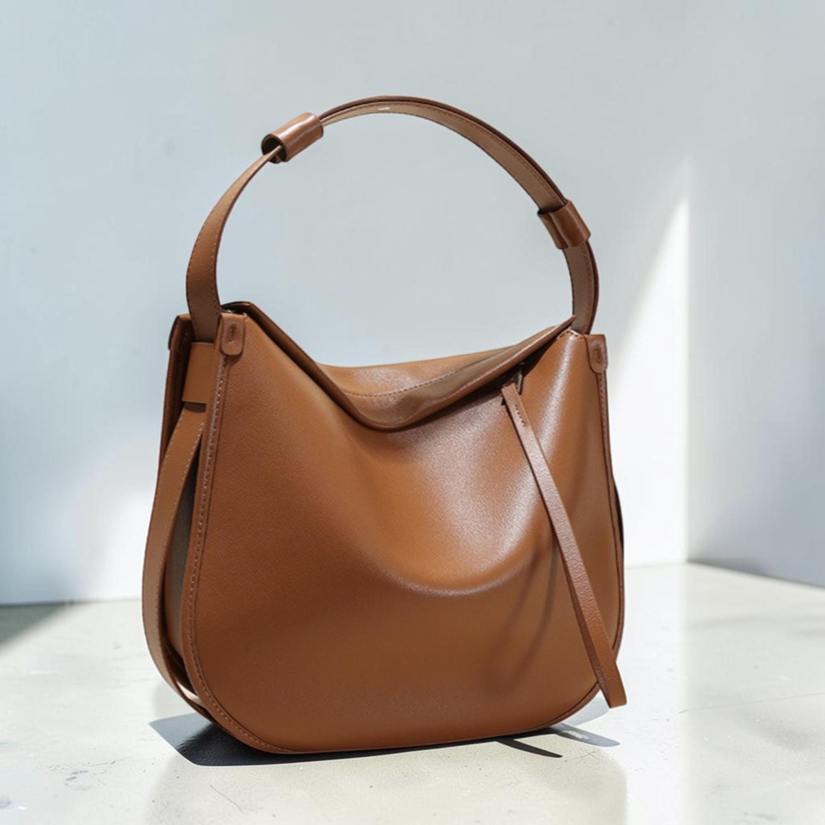 May include: A brown leather shoulder bag with a curved design. The bag has a single handle and a long strap detail on the side. The bag is made of smooth leather and has a minimalist design.