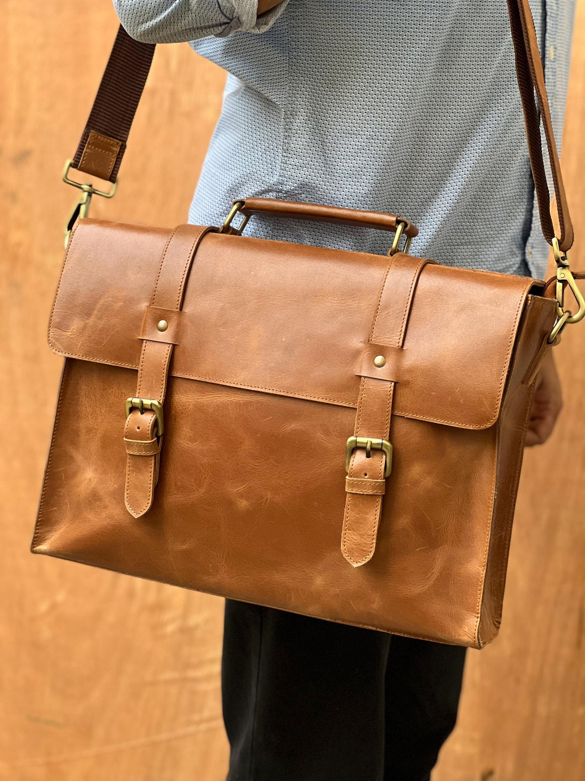 May include: A brown leather briefcase with a shoulder strap and a top handle. The briefcase has two front straps with buckles and brass-colored hardware. The bag is rectangular and appears to be made of high-quality leather.