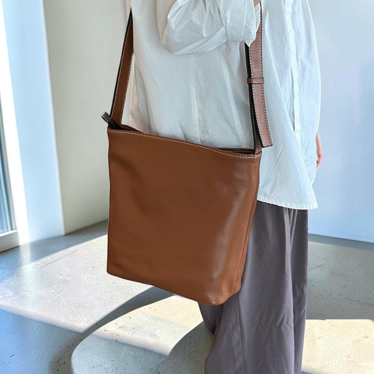 May include: A brown leather shoulder bag with a long strap. The bag is rectangular with a simple design and white stitching. The strap is brown with a black interior. The bag is worn over the shoulder.