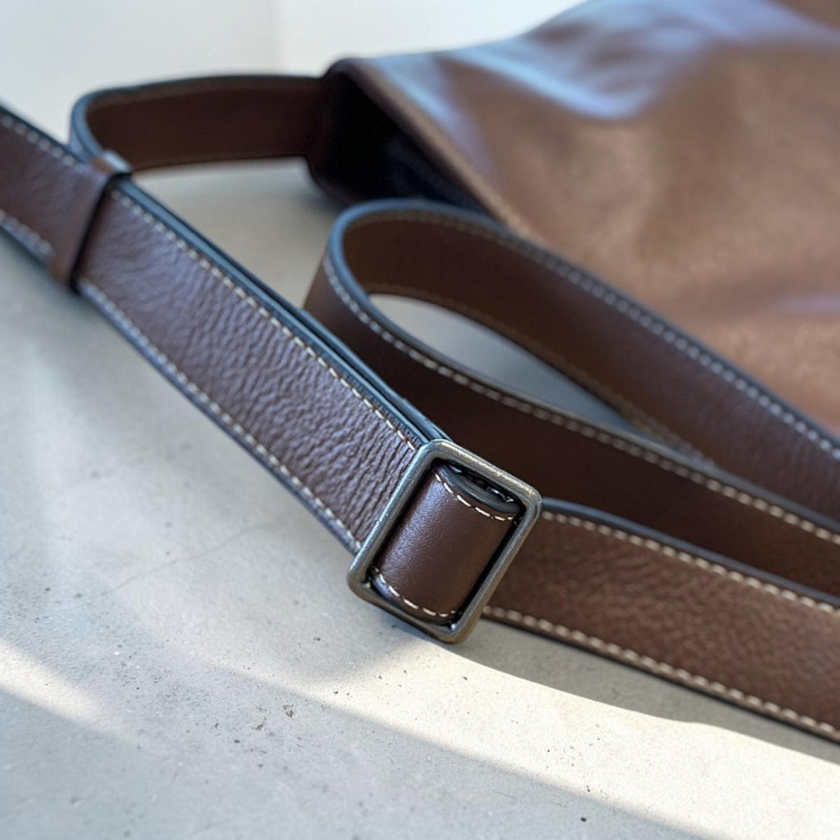 May include: Close-up of a brown leather shoulder bag with a matching adjustable strap. The strap features white stitching and a silver-colored metal buckle. The bag's body is visible in the background.