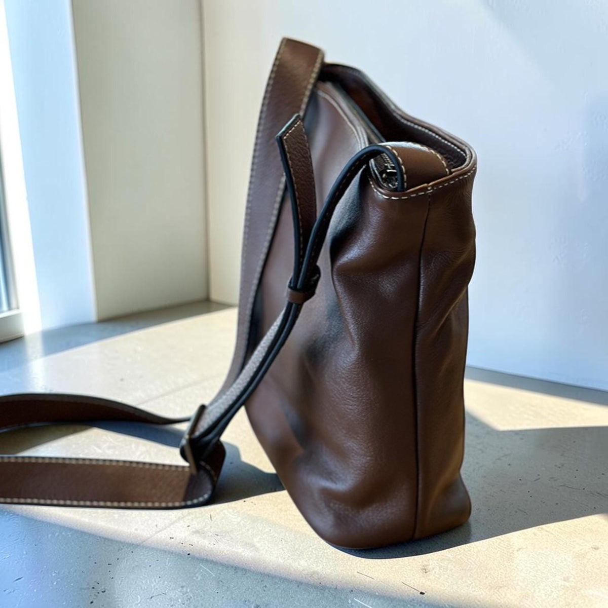 May include: A brown leather crossbody bag with a long adjustable strap. The bag features white stitching along the seams and a zipper closure. The bag is made of a soft, textured leather.