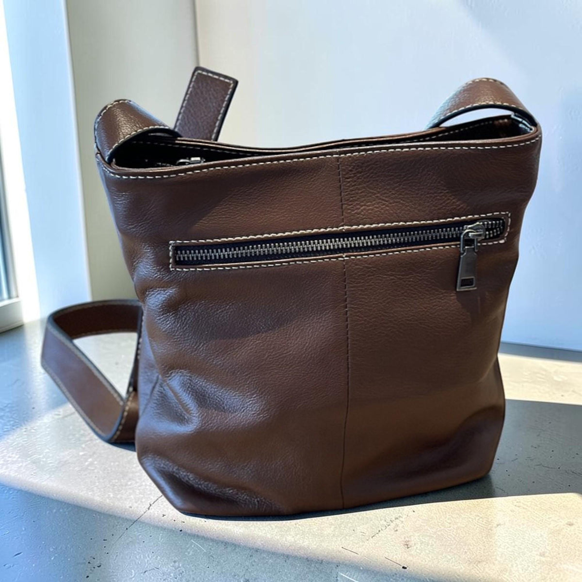 May include: A brown leather shoulder bag with a zip closure and a long strap. The bag has white stitching details and a small handle at the top. The bag is sitting on a light gray surface.