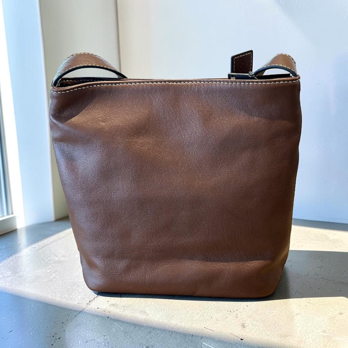 May include: A brown leather bucket bag with a single shoulder strap. The bag has a simple design with visible stitching and a small, rectangular closure. The bag is sitting on a light-colored surface.
